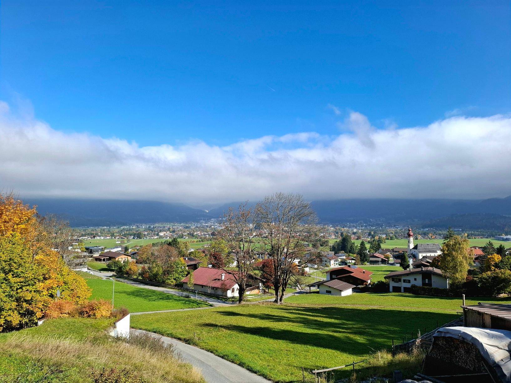 Herbstlicher Panoramablick über ein grünes Tal mit bunten Bäumen, Dorf und Bergen unter einem bewölkten Himmel.