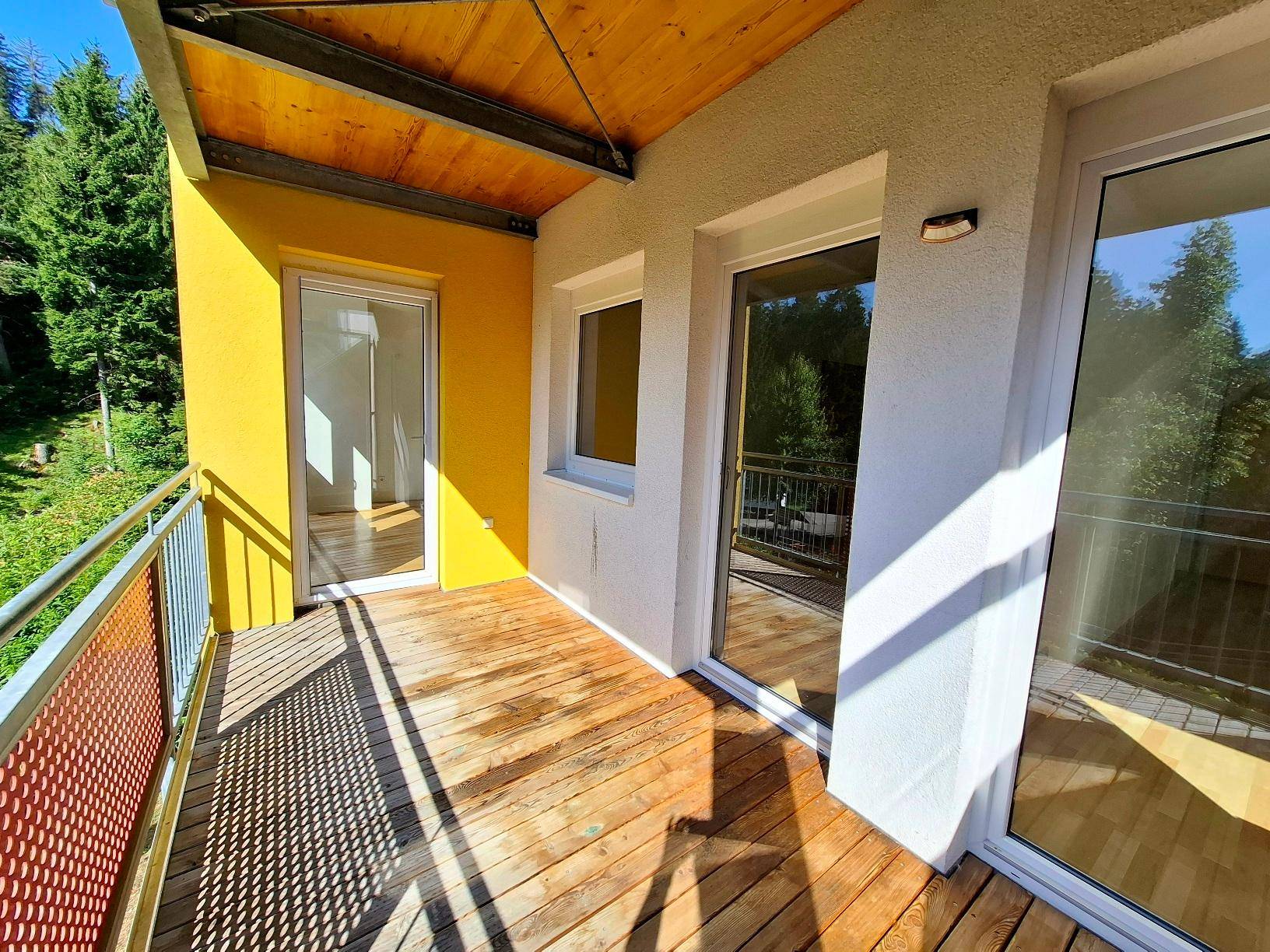 Geräumiger Balkon mit Holzboden und gelber Wand, umgeben von grünen Bäumen und Natur.