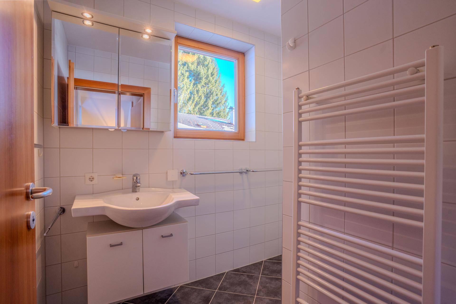 Modernes Badezimmer mit weißen Wandfliesen, dunklem Fliesenboden, Einzelwaschbecken und Spiegelschrank.