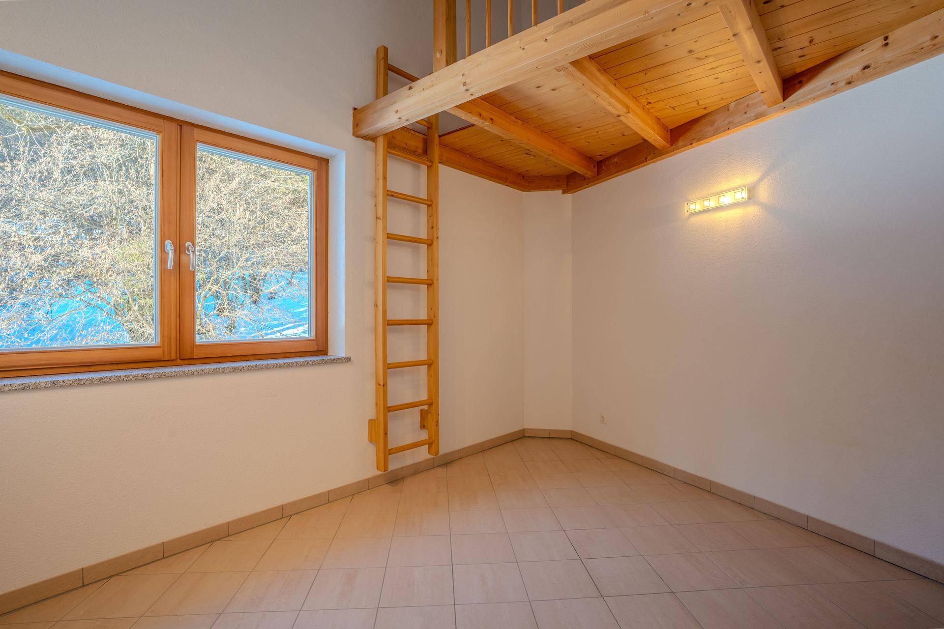Kleiner Raum mit Fliesenboden, Fenster und einer Holzleiter, die zu einem gemütlichen Schlafbereich führt.
