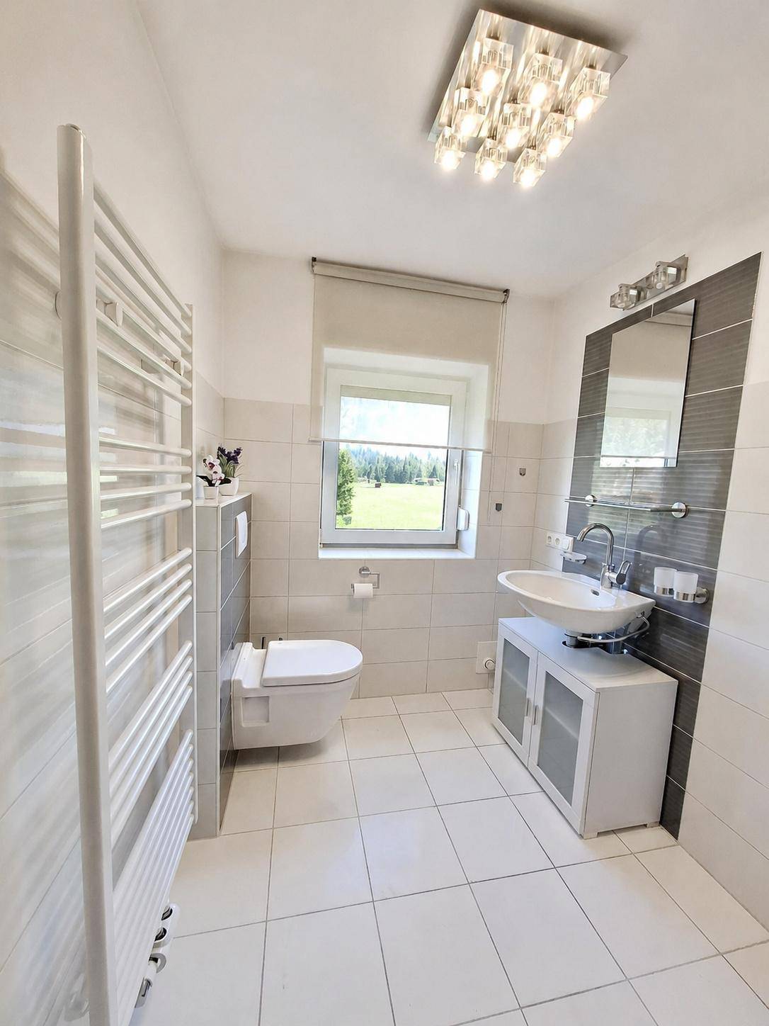 Modernes Badezimmer mit weißen und grauen Fliesen, wandhängendem WC und Fenster mit Grünblick.