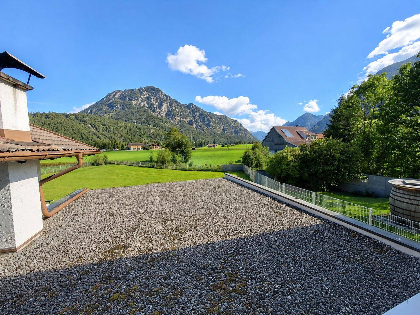 Weitläufiger Blick von einer Terrasse über grüne Felder und majestätische Berge unter blauem Himmel.