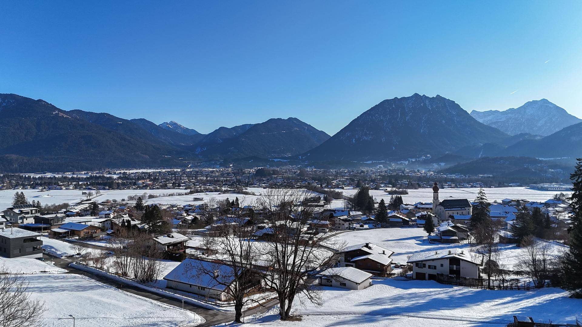 Winterlicher Panoramablick über ein schneebedecktes Tal mit Dorf und imposanten Bergen unter blauem Himmel.
