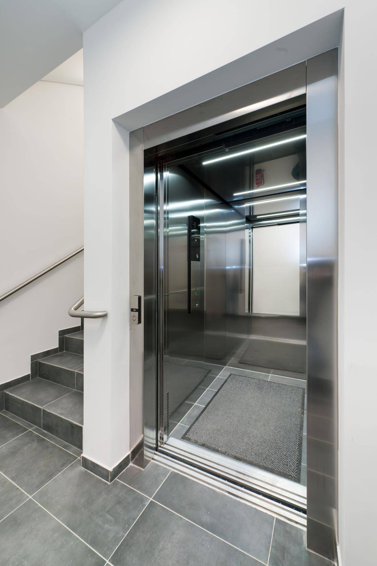 Moderner Aufzug mit Edelstahloberflächen und angrenzendem Treppenhaus im Gebäude.