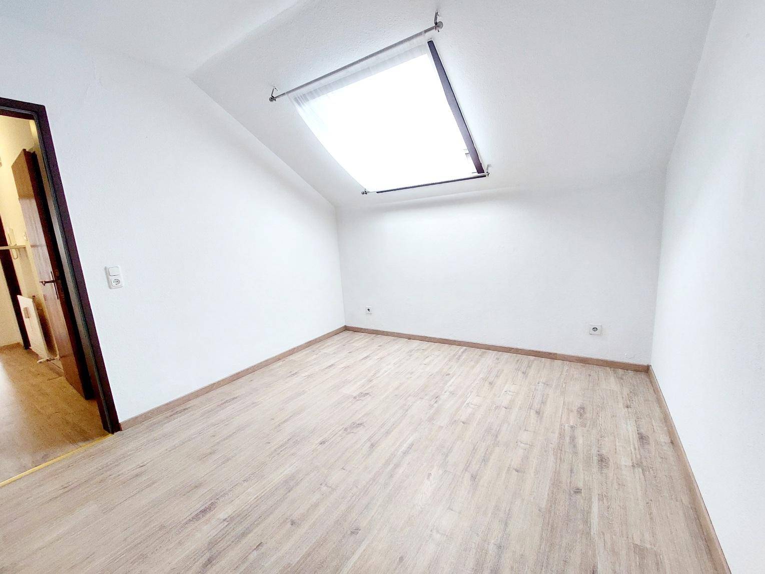 Leerer Raum im Dachgeschoss mit hellem Laminatboden, Schrägdecke und einem Dachfenster.
