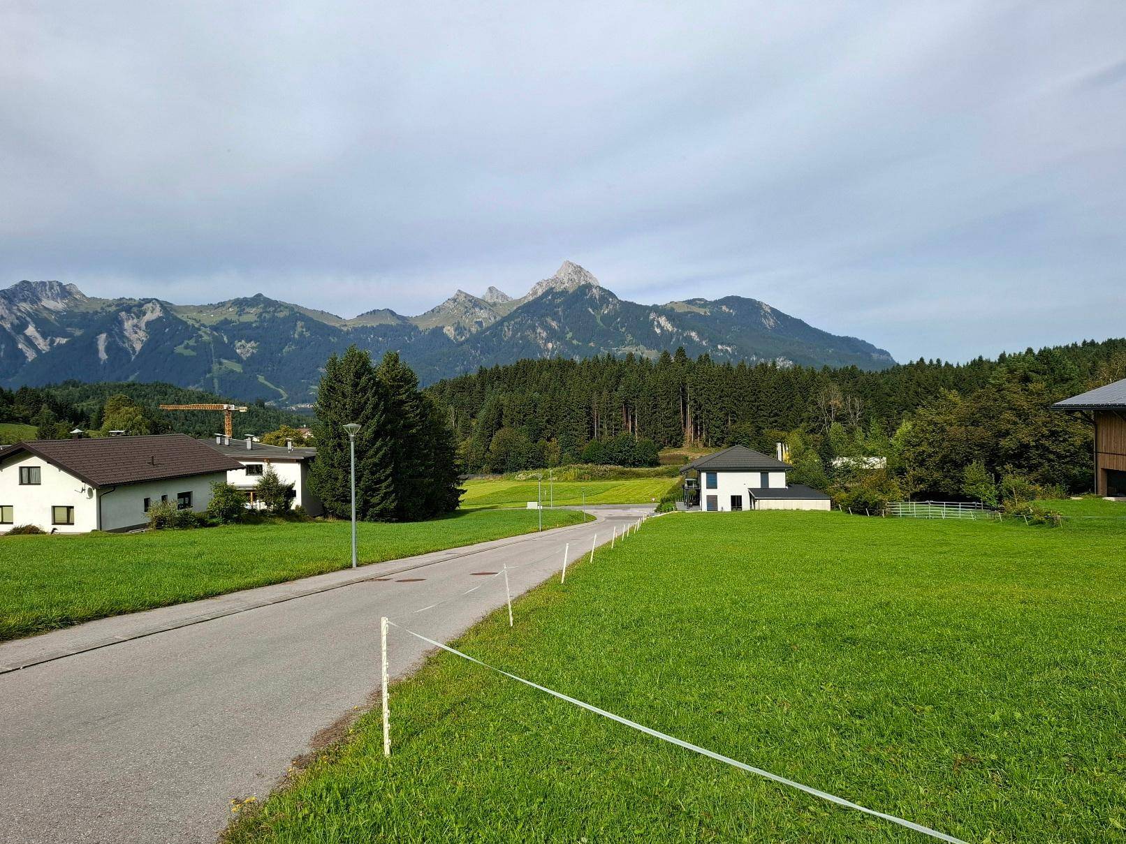 Weitläufige Landschaft mit Wohnhäusern, saftigen Wiesen und majestätischem Alpenpanorama im Hintergrund.