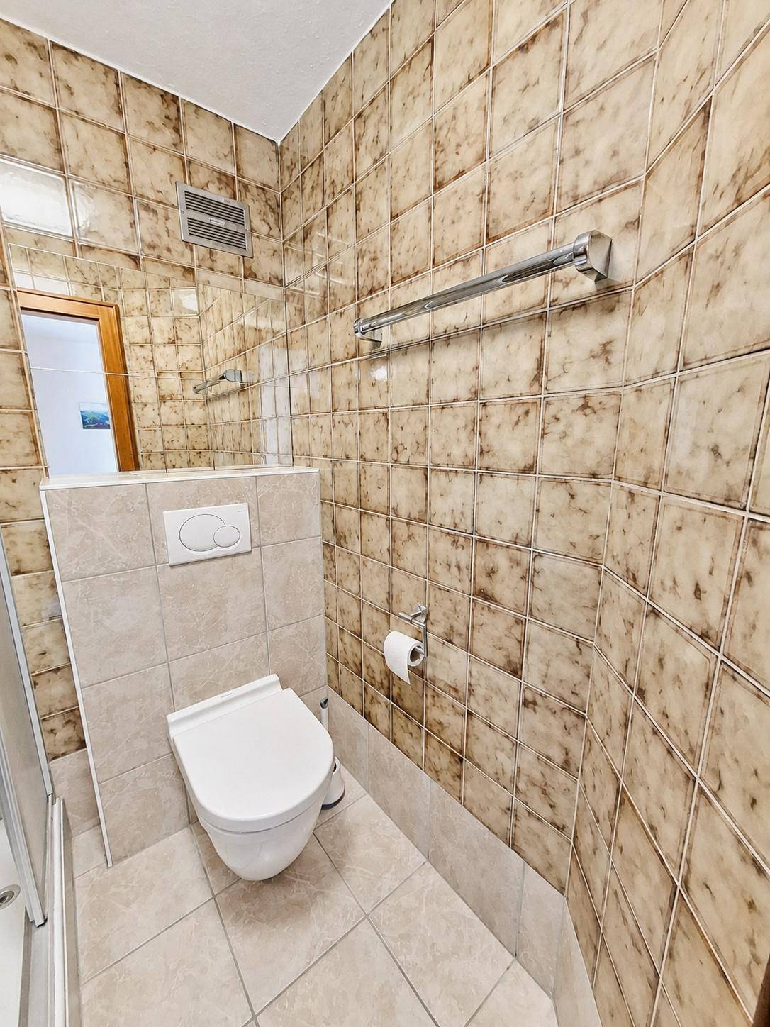 Badezimmer mit braunen Fliesen, wandhängendem WC und einem Spiegel über dem Waschbecken.