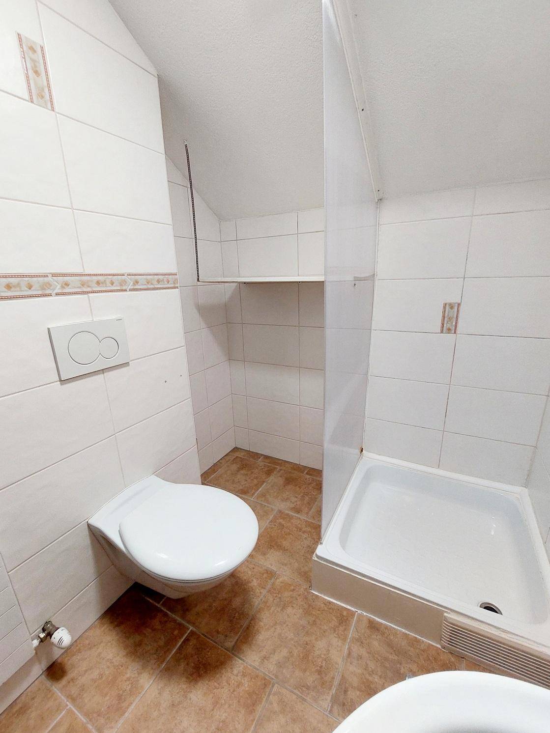 Detailansicht des Badezimmers mit wandhängender Toilette und Duschbereich mit Fliesen.