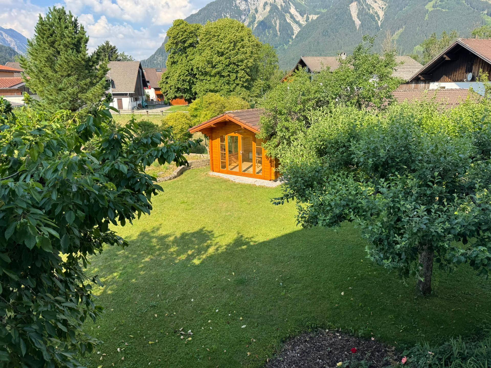 Idyllischer Garten mit einem Holzgartenhaus, umgeben von Bäumen und Sträuchern mit Bergblick.
