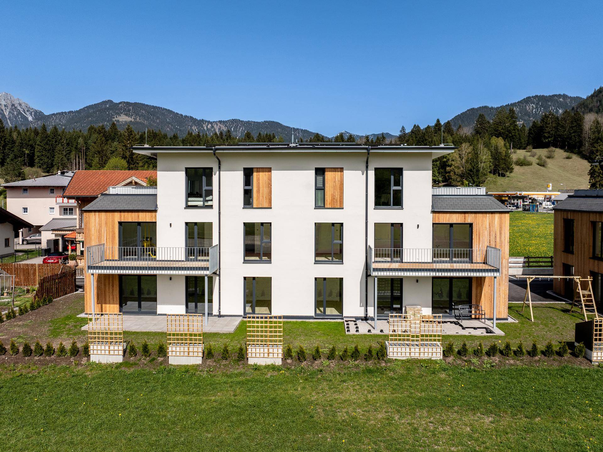 Frontansicht des Neubaus mit Balkonen und Holzverkleidung, umgeben von grüner Landschaft.
