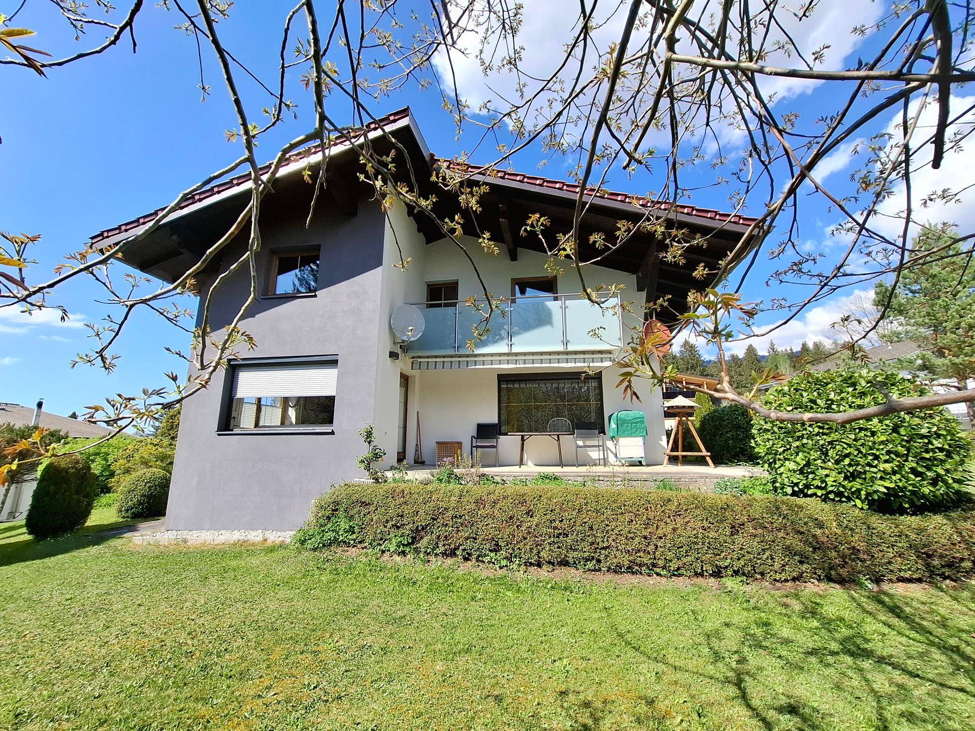 Wohnhaus mit Terrasse und Balkon, umgeben von gepflegtem Garten und Natur unter strahlendem Himmel.