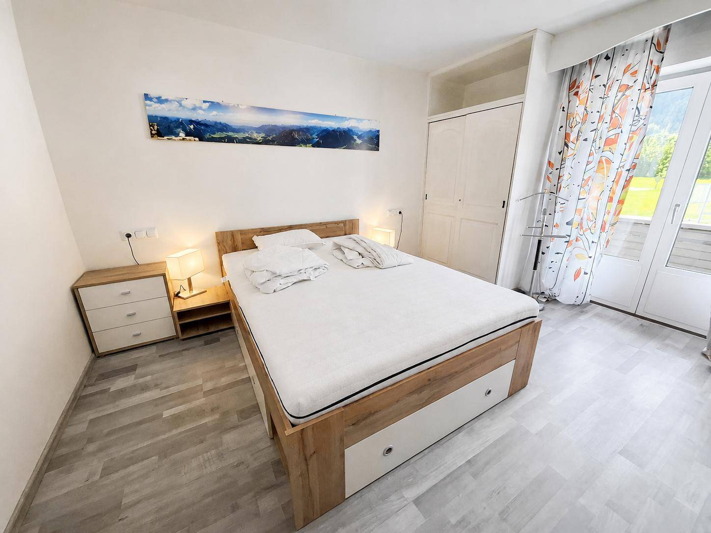 Helles Schlafzimmer mit Holzbett, Nachttischen, weißem Kleiderschrank und Zugang zu einem Außenbereich.