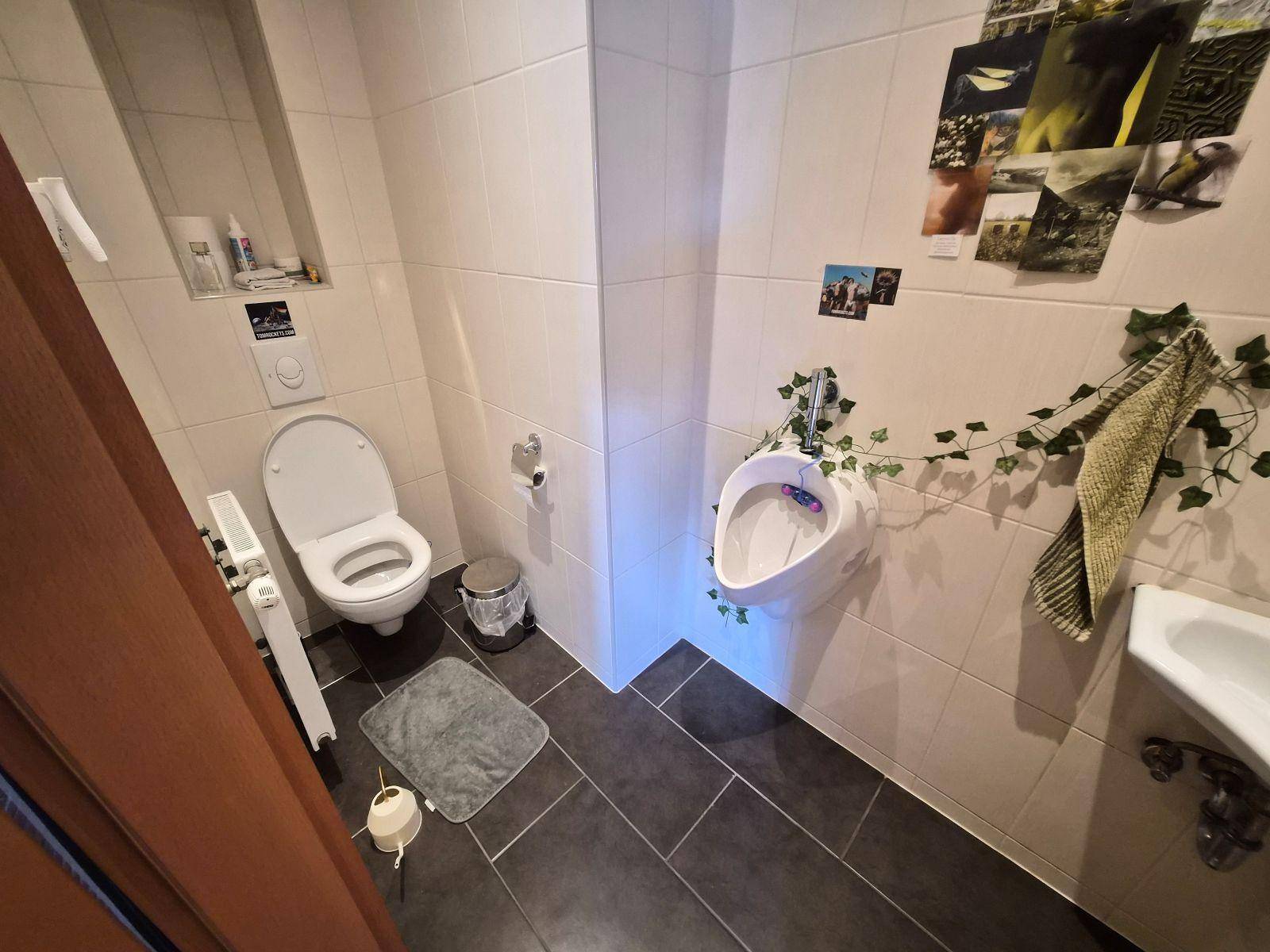 Separate Toilette mit WC, Urinal, kleinem Waschbecken und Fliesenboden.