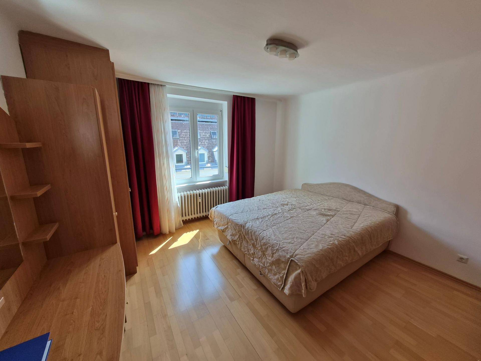 Helles Schlafzimmer mit Doppelbett, großem Holzschrank und Fenster mit roten Vorhängen.