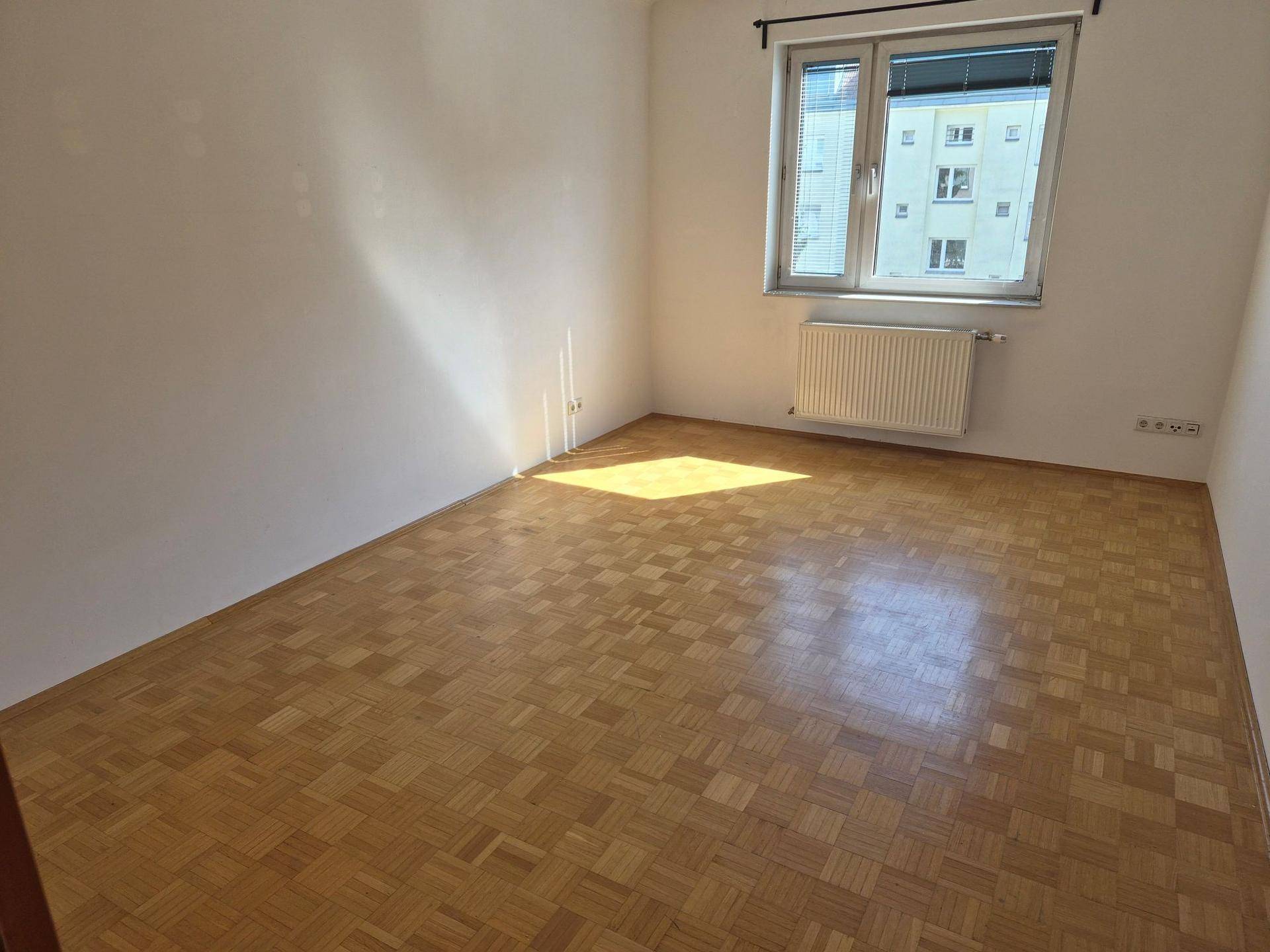Helles, leeres Zimmer mit Parkettboden, weißen Wänden und viel natürlichem Licht.