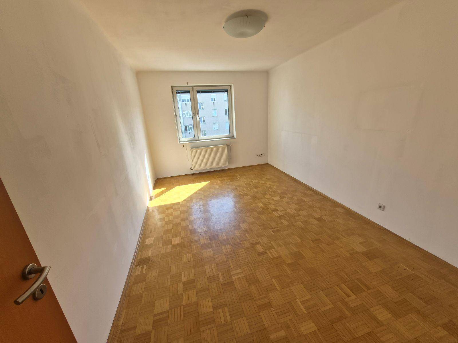 Sonniger, leerer Raum mit Parkettboden, weißen Wänden und einem Fenster.