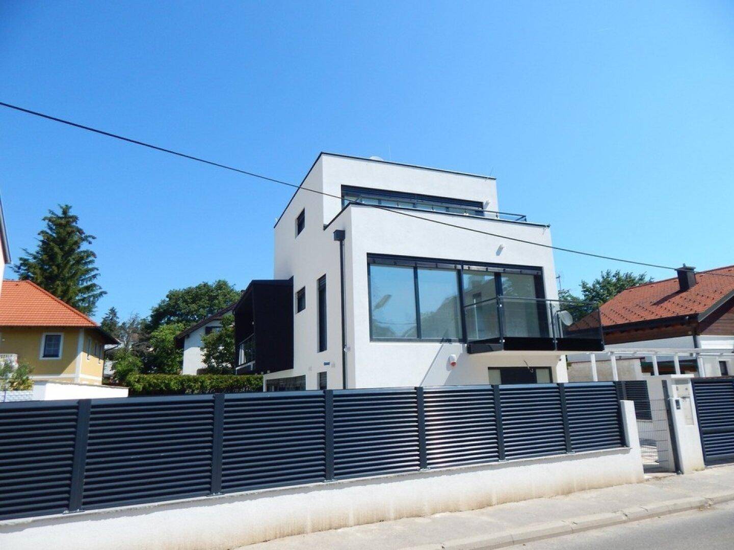 Moderne weiße Villa mit großen Fenstern und Flachdach, umgeben von einem dunklen Zaun.