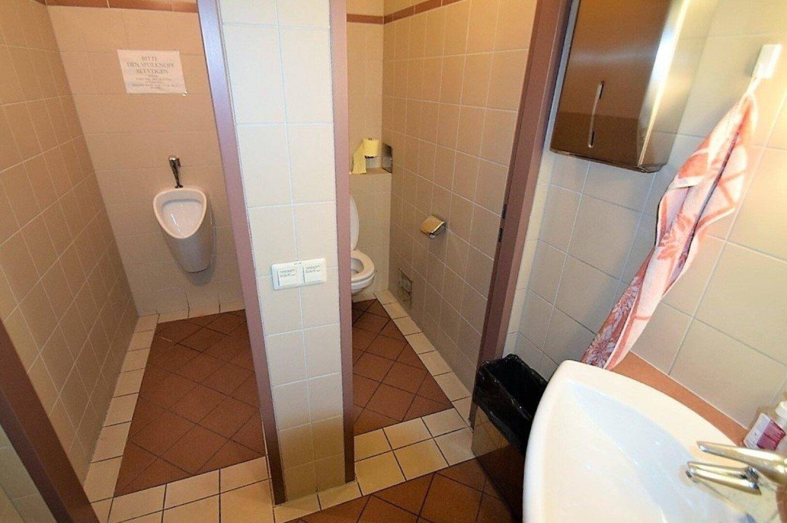 Gefliester Sanitärbereich mit Urinal, Toilette in einer Kabine und Waschbecken, funktional und sauber.