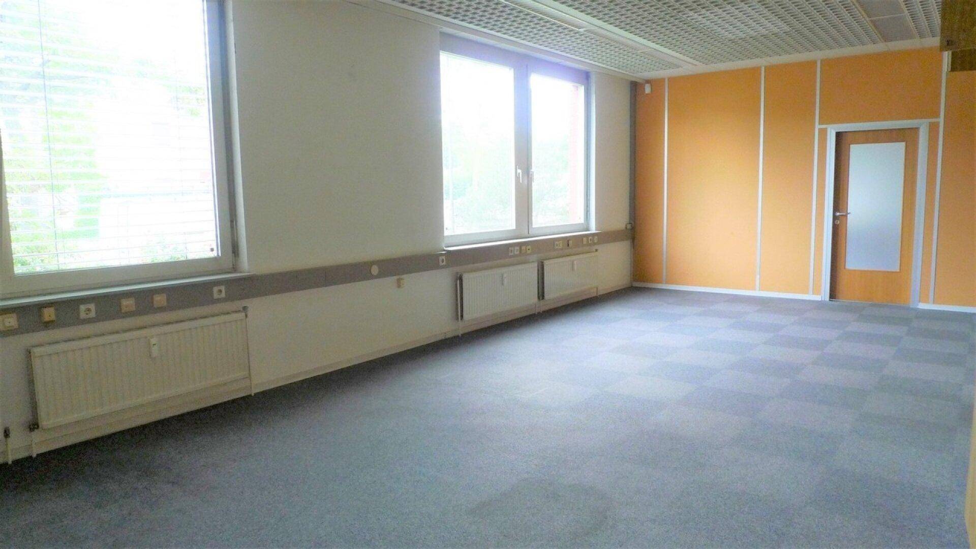 Geräumiges Büro mit großen Fenstern, Teppichboden und orangefarbenen Wandpaneelen, bereit zur individuellen Gestaltung.