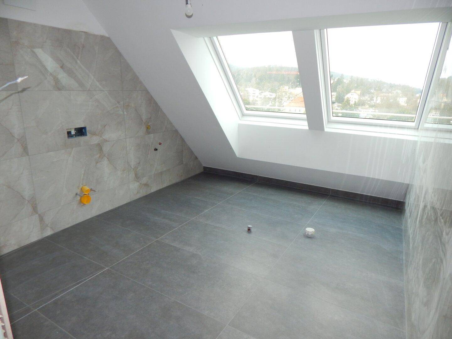 Badezimmer im Dachgeschoss mit grauen Bodenfliesen, Marmoroptik-Wandfliesen und Dachfenstern.