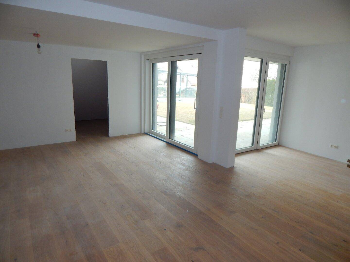Unfertiger Wohnbereich mit Holzboden und großen Fenstern, die viel Tageslicht hereinlassen.