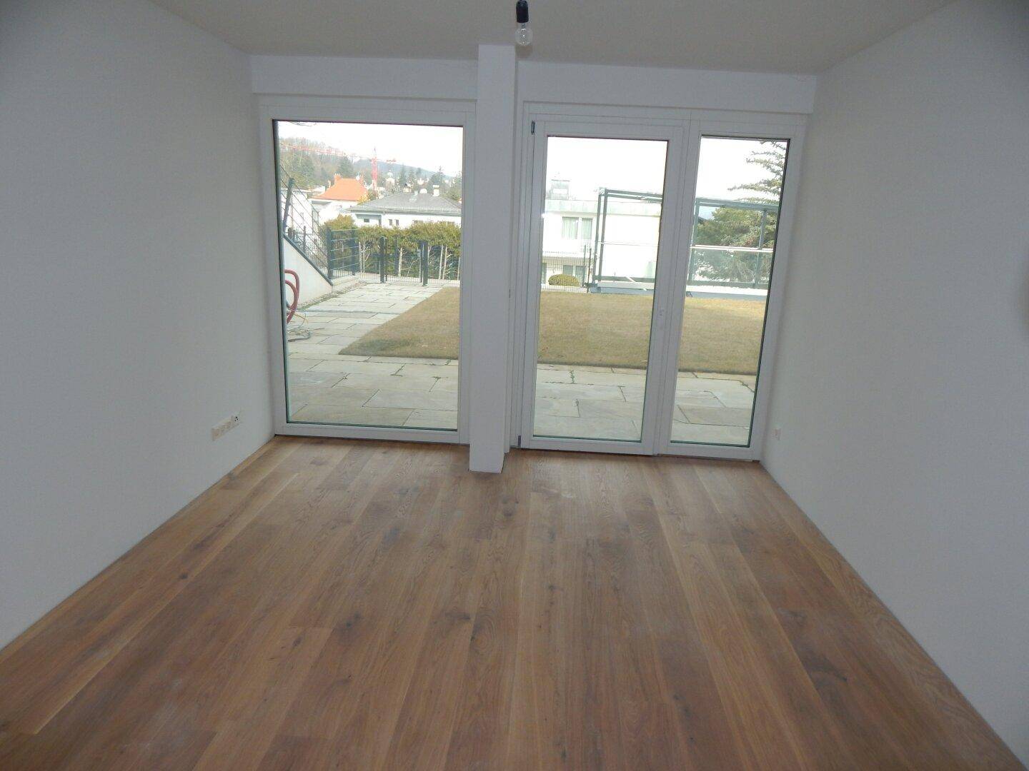 Heller Raum mit Holzboden und großen Fenstern, die den Blick auf den Außenbereich freigeben.
