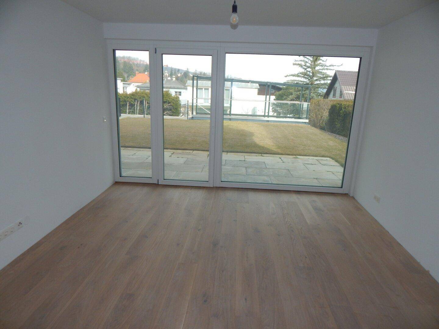 Unfertiger Raum mit Holzboden und breiter Fensterfront mit Blick auf den Gartenbereich.