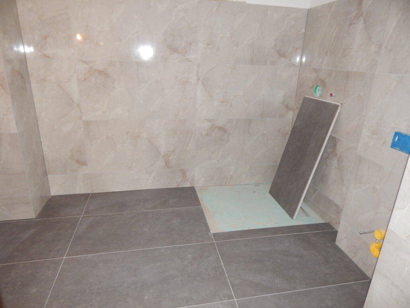 Unfertiges Badezimmer mit grauen Bodenfliesen und hellen Marmoroptik-Wandfliesen.
