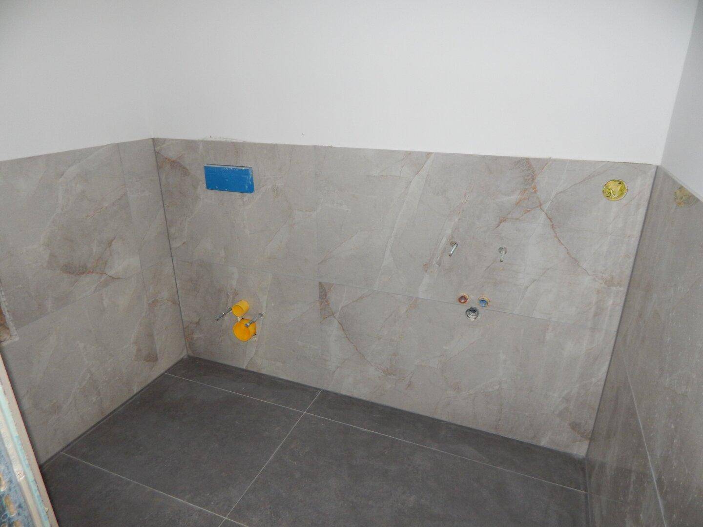Unfertiger kleiner Raum mit Fliesen an Wänden und Boden, vorbereitet für Sanitärinstallationen.