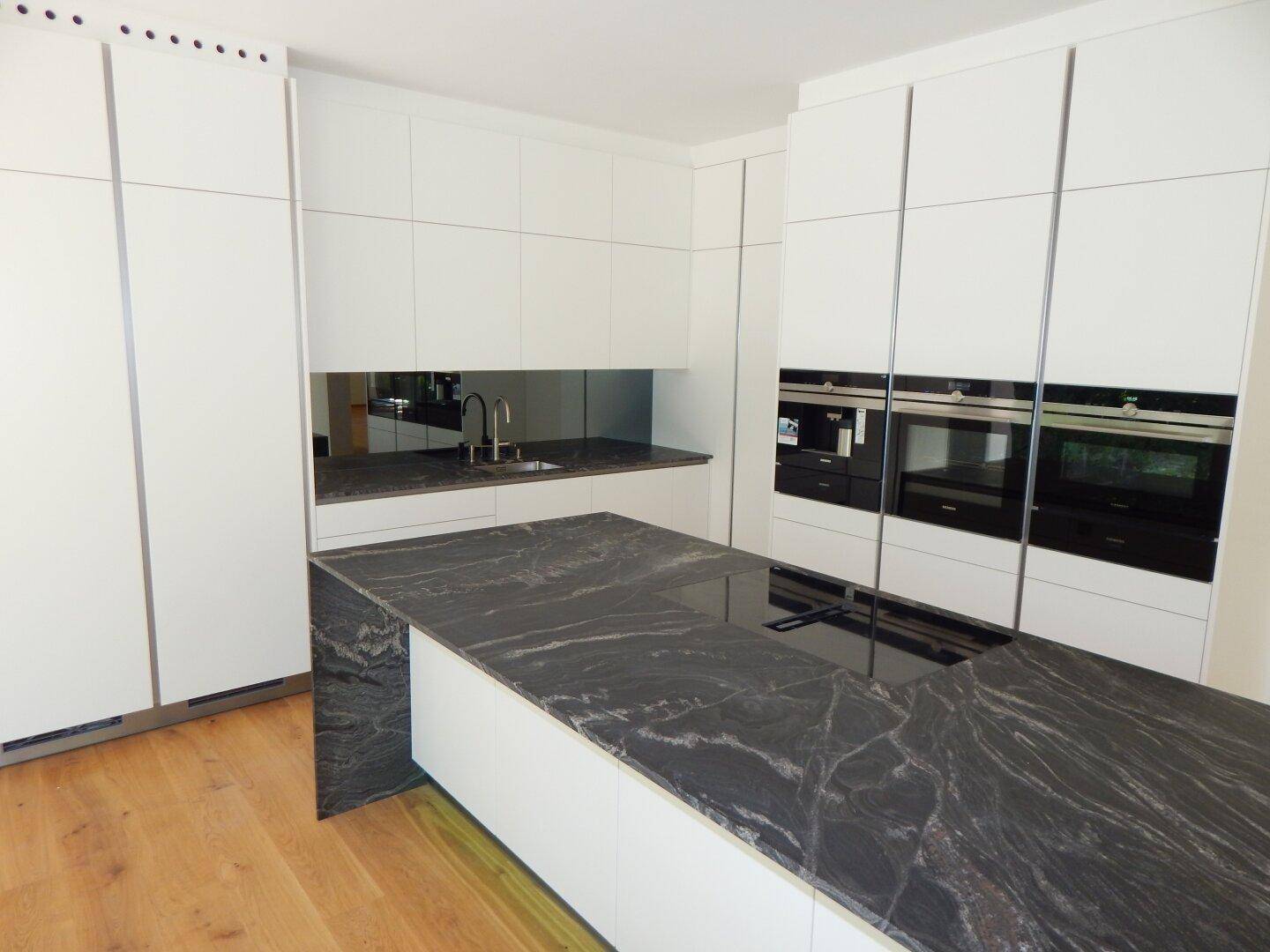 Moderne Küche mit weißer Schrankfront, dunkler Steininsel und integrierten Geräten.