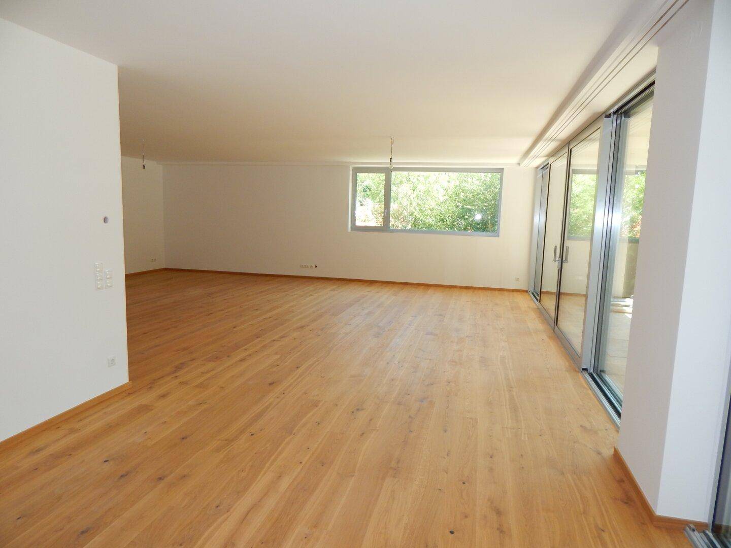 Heller, leerer Wohnraum mit Holzparkett und großen Fenstern, die viel Tageslicht hereinlassen.