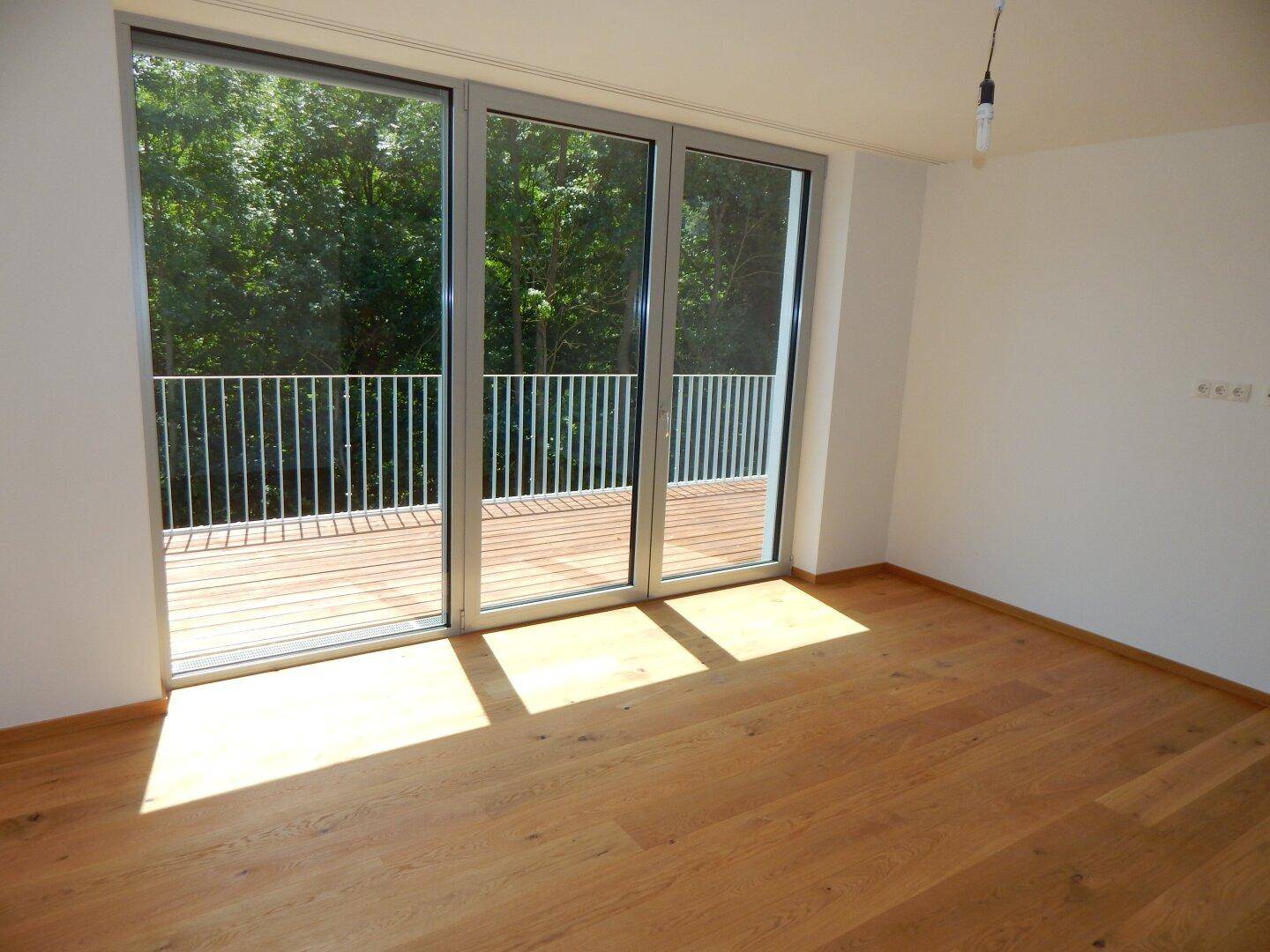 Lichtdurchflutetes Zimmer mit Holzboden und breiter Fensterfront zum Außenbereich.