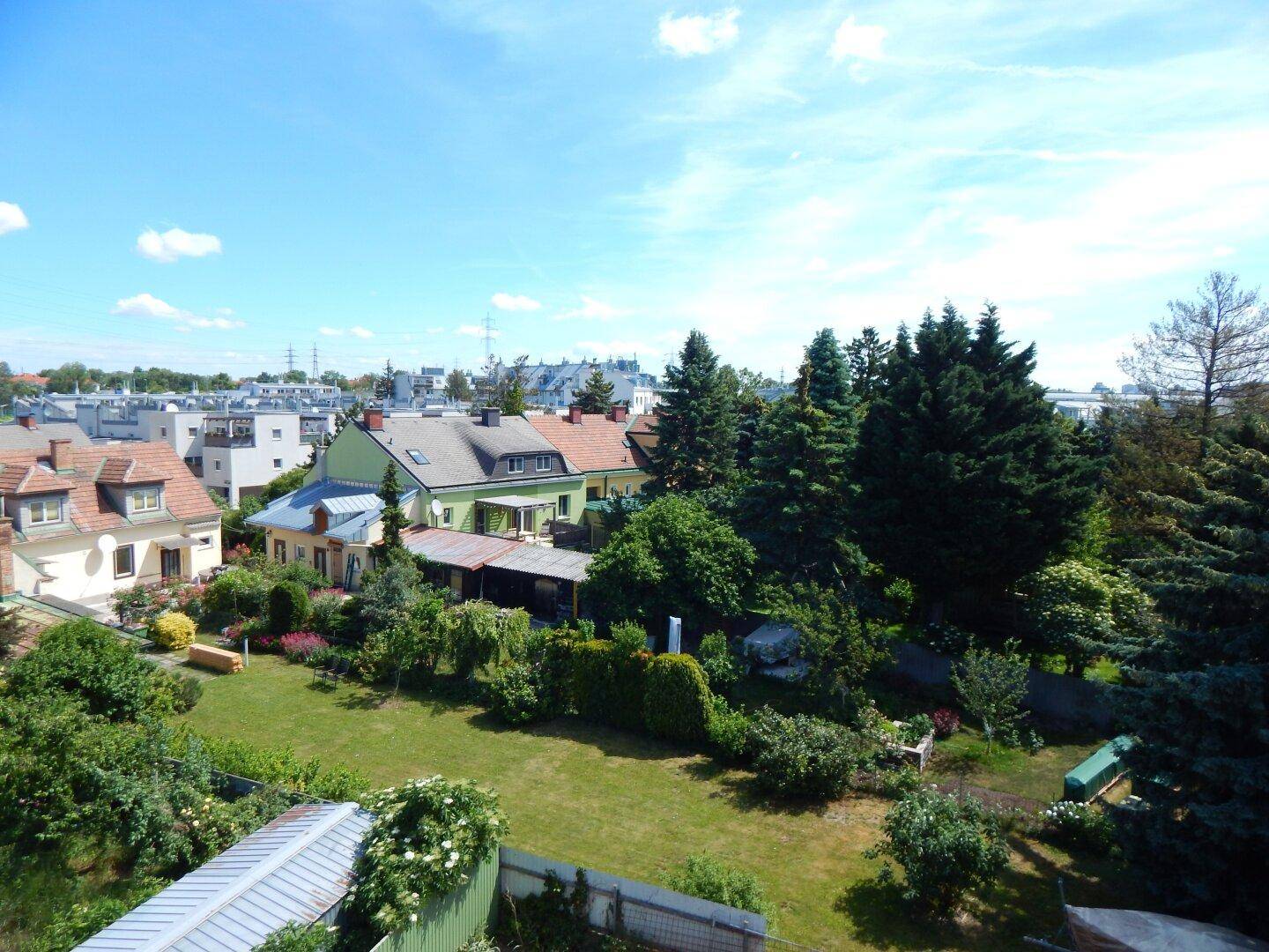 Panoramablick über eine Wohnsiedlung mit gepflegten Gärten, alten Bäumen und traditionellen Häusern unter blauem Himmel.