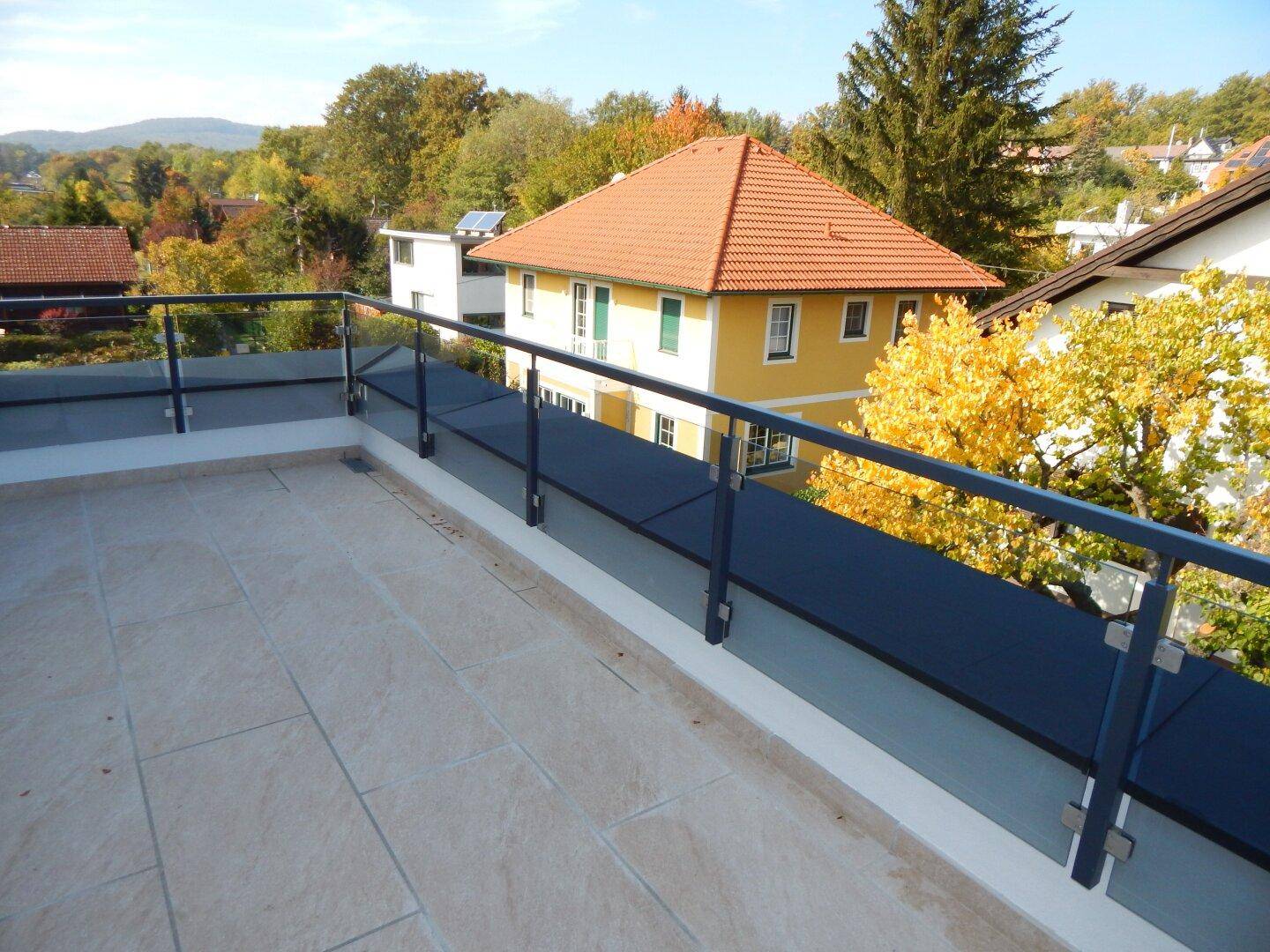 Heller Balkon mit Glasbrüstung und weitem Blick auf die grüne Umgebung und Nachbarhäuser.