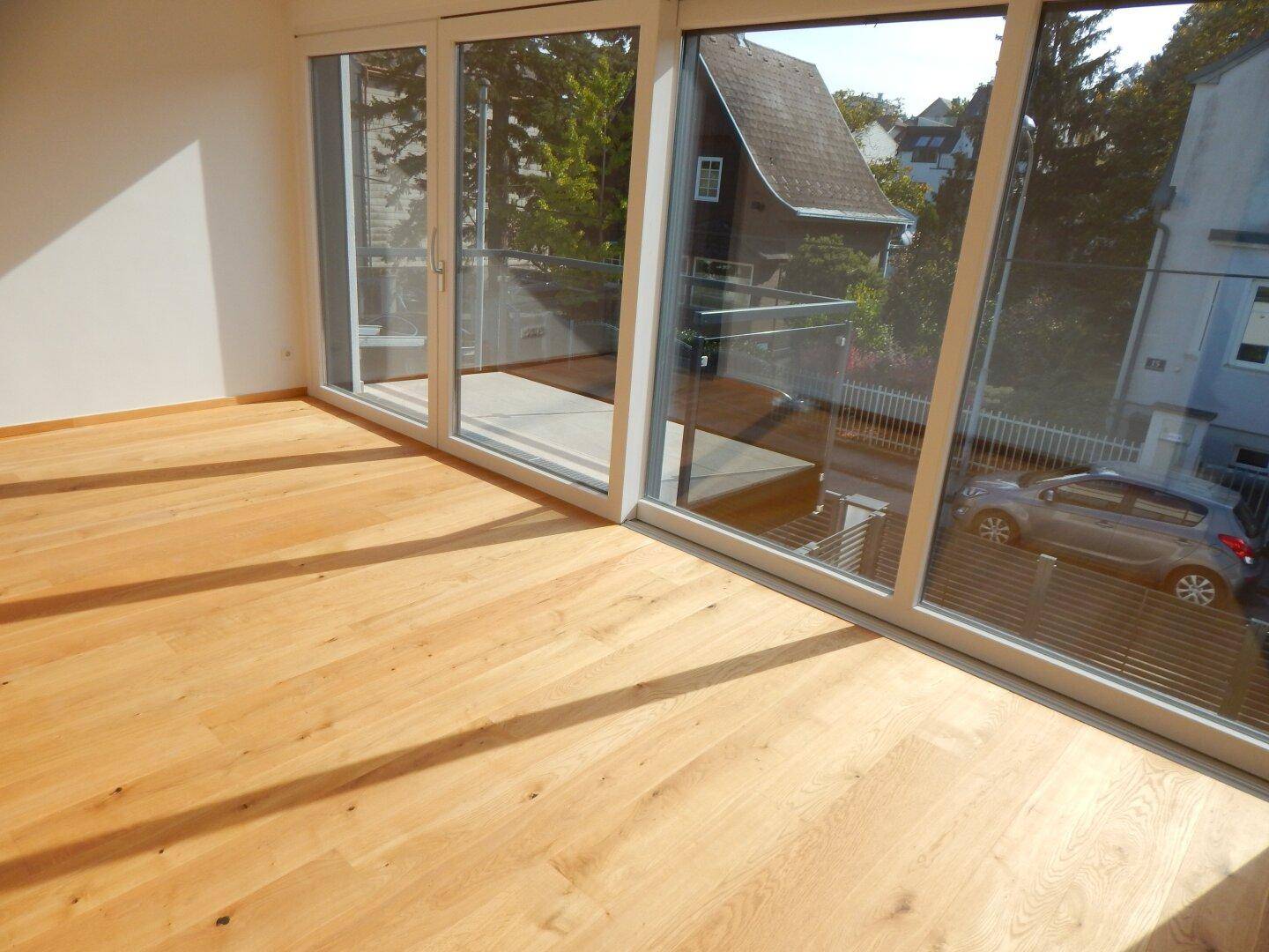 Heller Raum mit Holzparkett und großen Fenstern, die Zugang zu einem Balkon bieten.