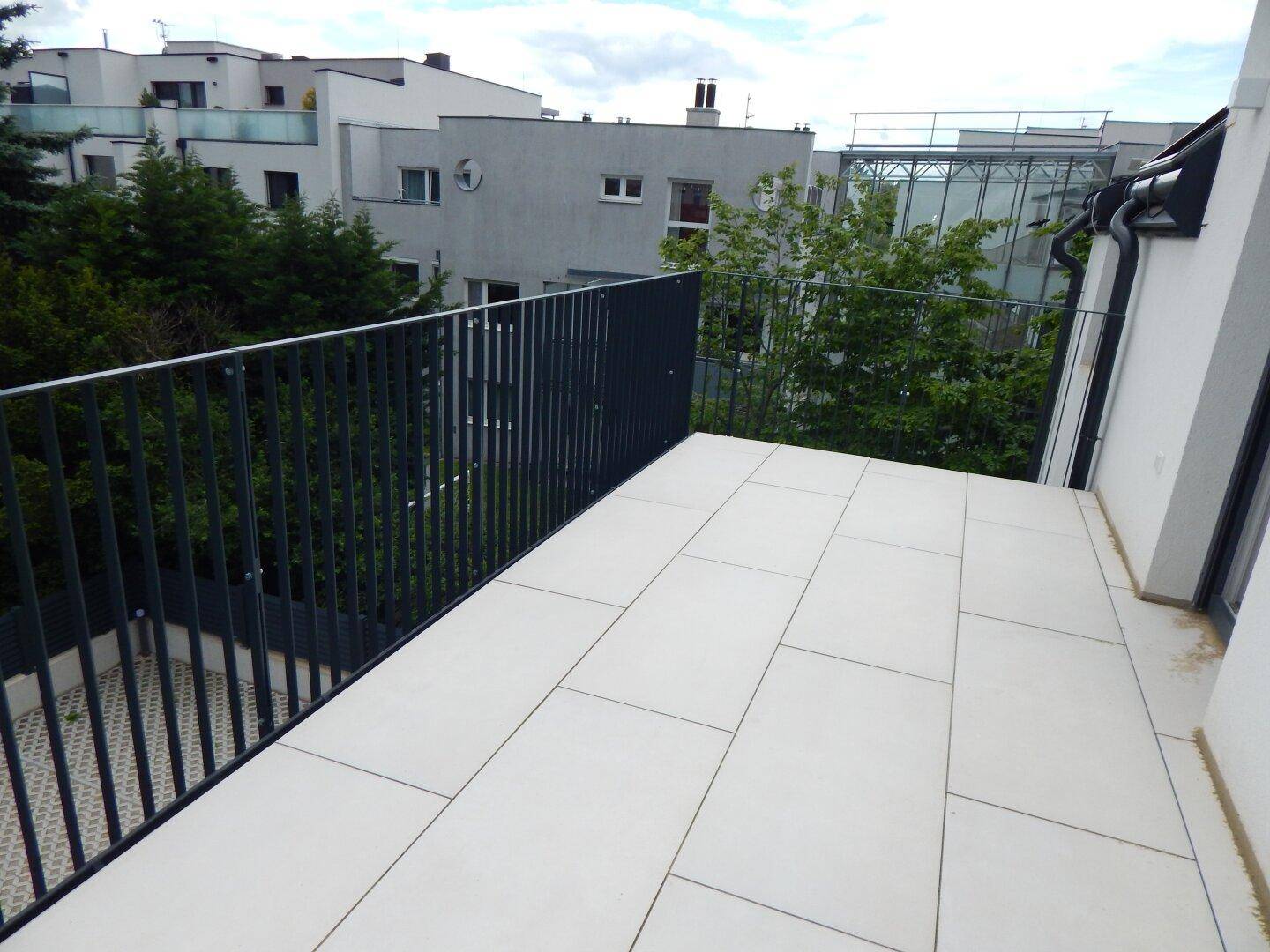 Moderne Terrasse mit hellen Fliesen und dunklem Geländer, umgeben von Bäumen und anderen Gebäuden.