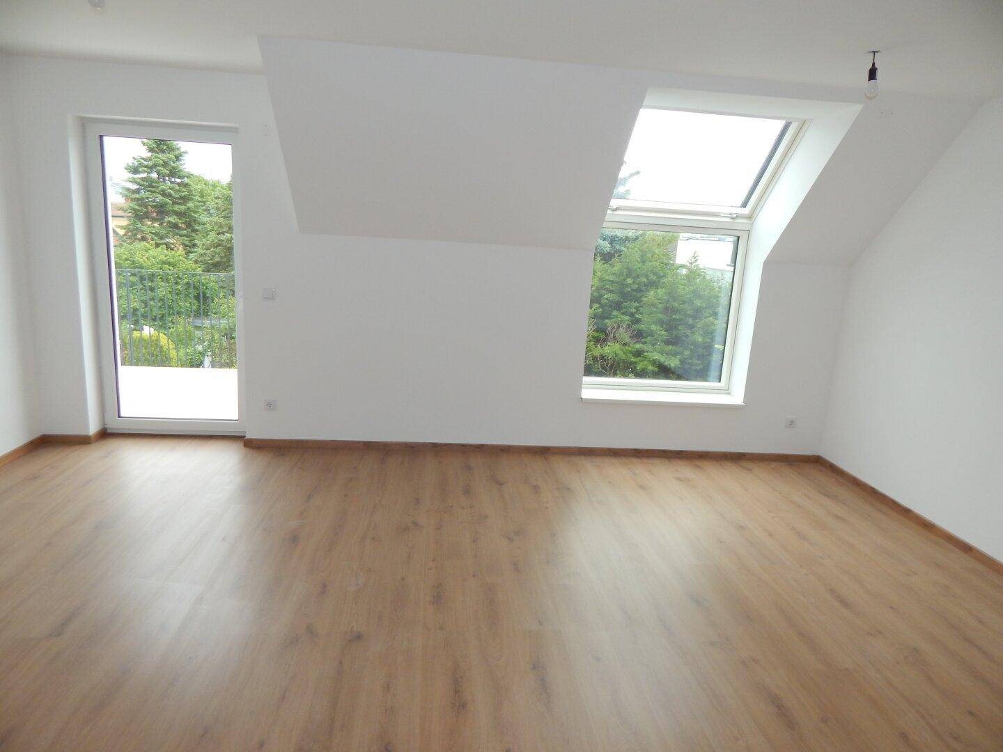 Geräumiger, heller Raum mit Laminatboden, Dachfenster und Balkontür, umgeben von viel Grün.