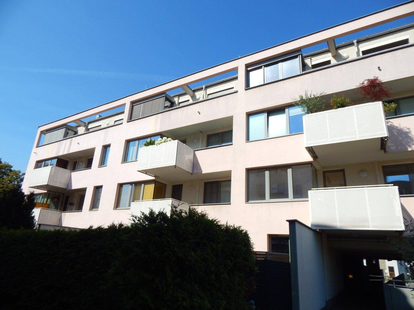Modernes Mehrfamilienhaus mit mehreren Balkonen und Tiefgaragenzufahrt unter blauem Himmel.