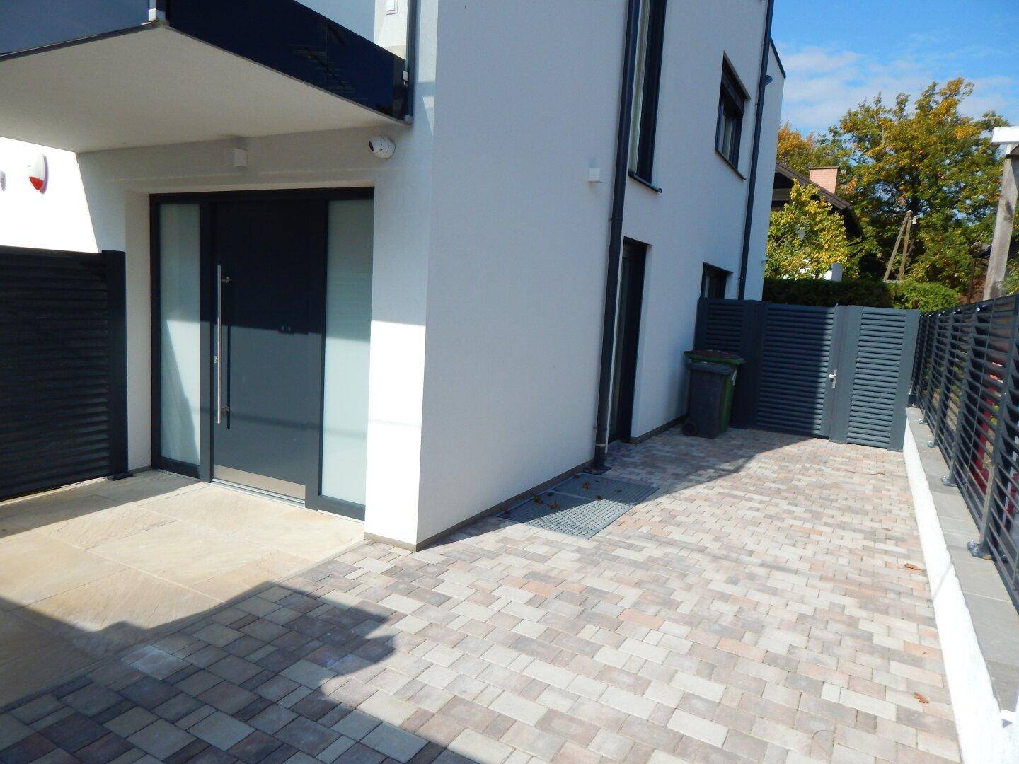 Moderner Hauseingang mit gepflastertem Vorplatz und dunklem Zaun.