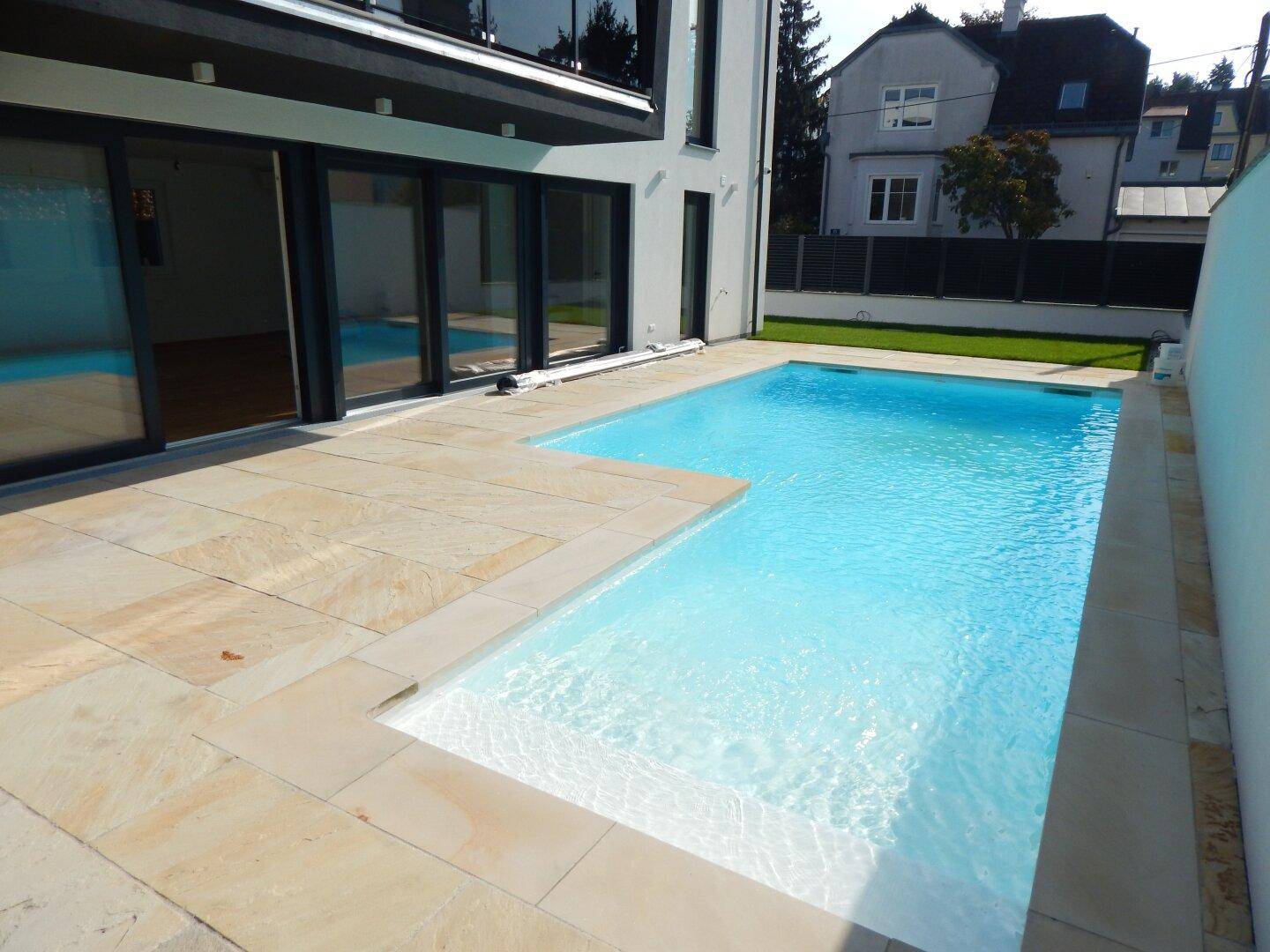 Großer, sauberer Poolbereich mit heller Steinterrasse und moderner Hausfassade.