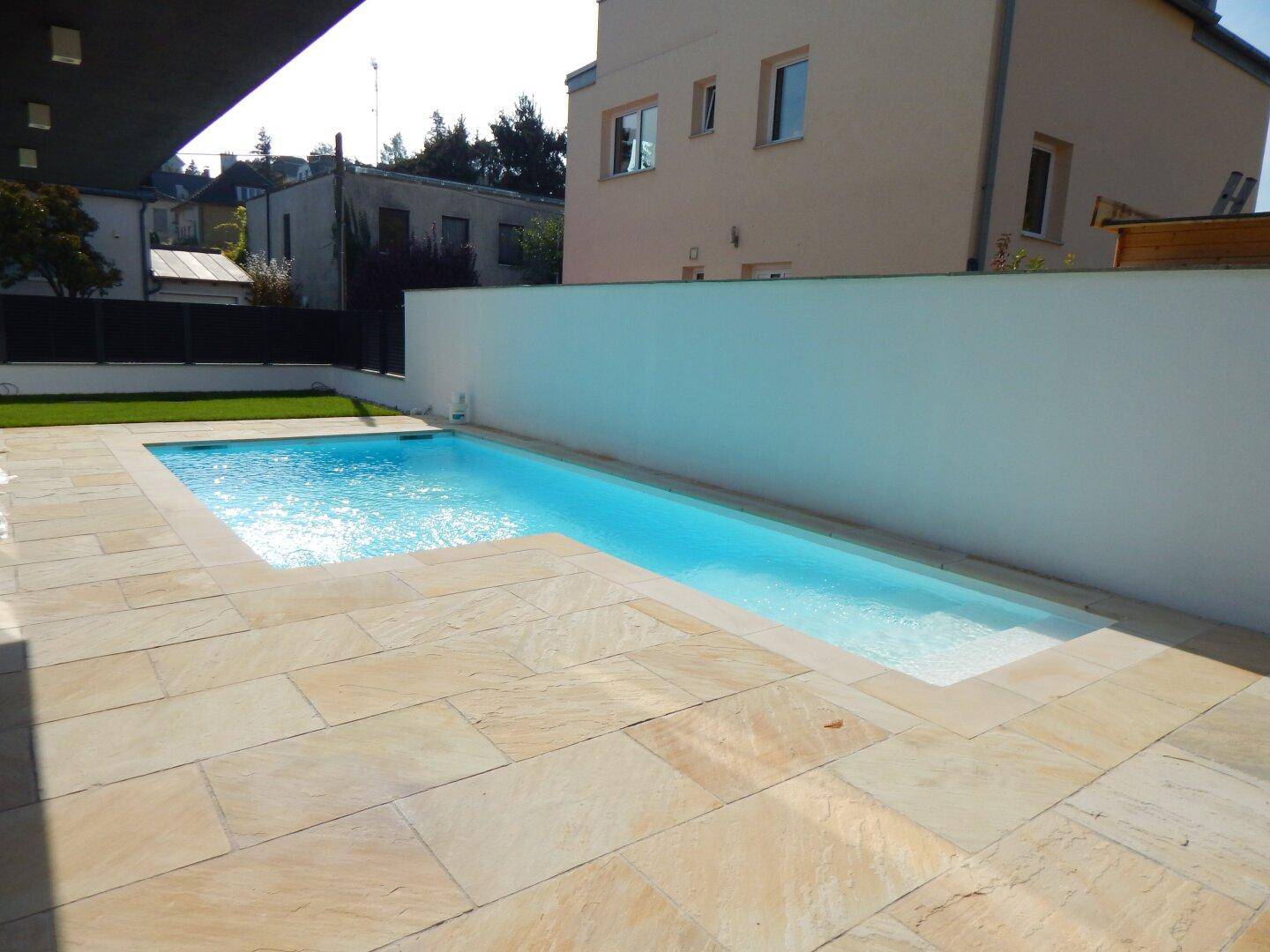 Schmaler Swimmingpool mit eleganter Steinterrasse und hoher weißer Gartenmauer.