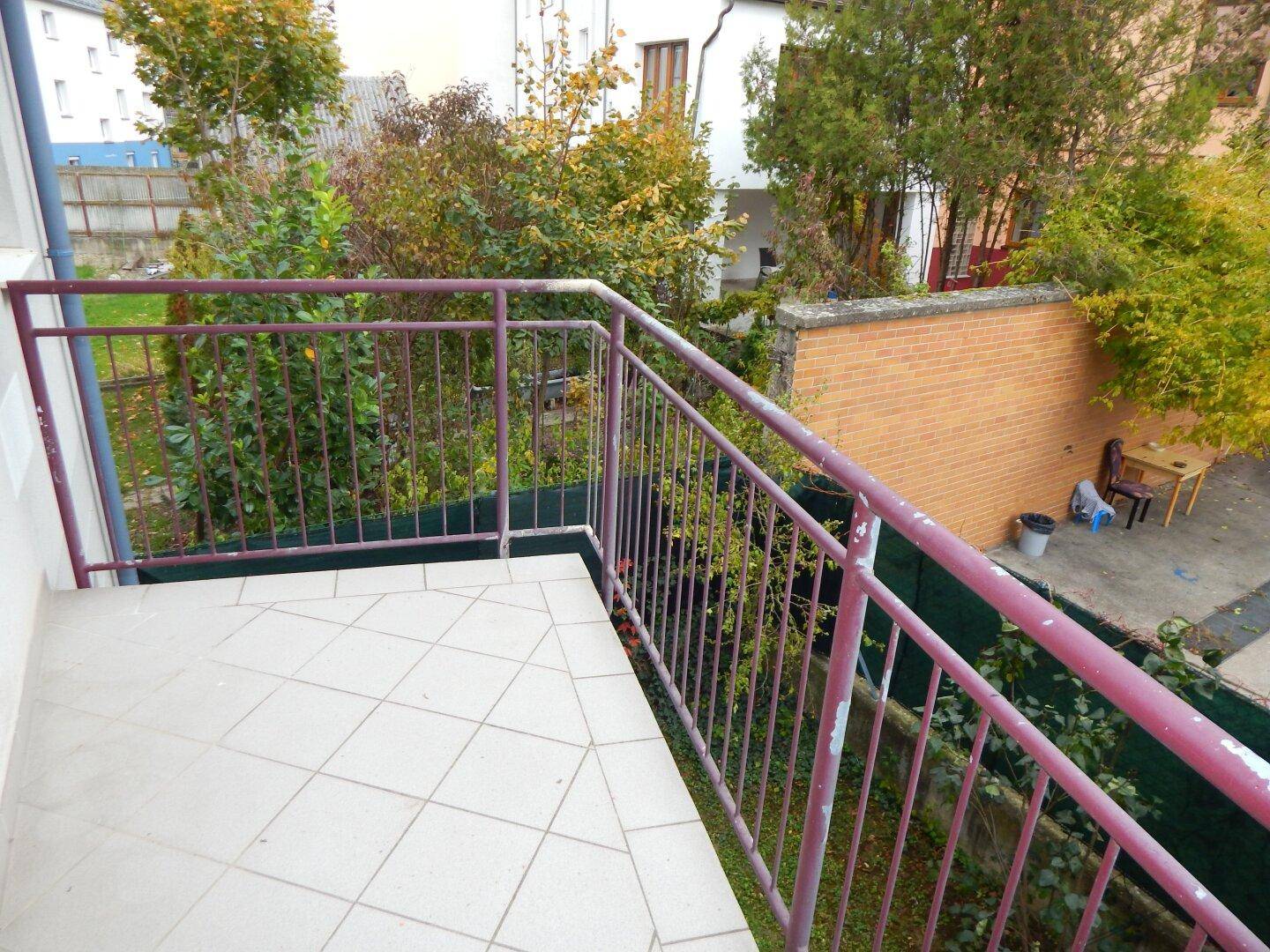 Balkon mit Fliesenboden und Blick auf Bäume und eine Ziegelmauer.