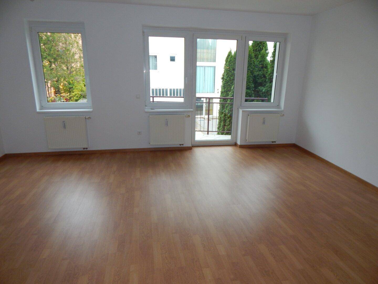 Heller Wohnraum mit Laminatboden und Zugang zu einem kleinen Balkon mit Blick auf Bäume.