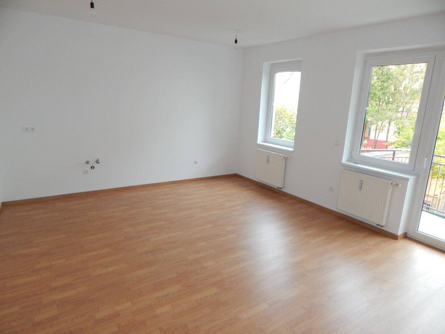 Leeres Wohnzimmer mit Laminatboden, weißen Wänden und Heizkörpern unter den Fenstern.