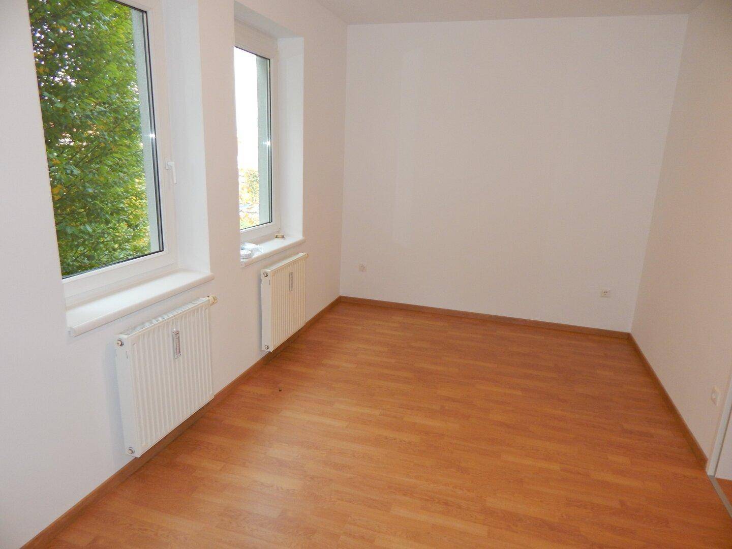 Leeres Zimmer mit Laminatboden und zwei Fenstern, bietet viel Tageslicht.
