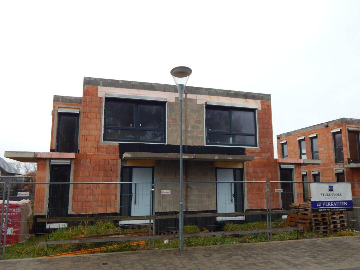 Fassade eines im Bau befindlichen Hauses mit großen Fenstern und Rollläden, gesehen von der Straße.