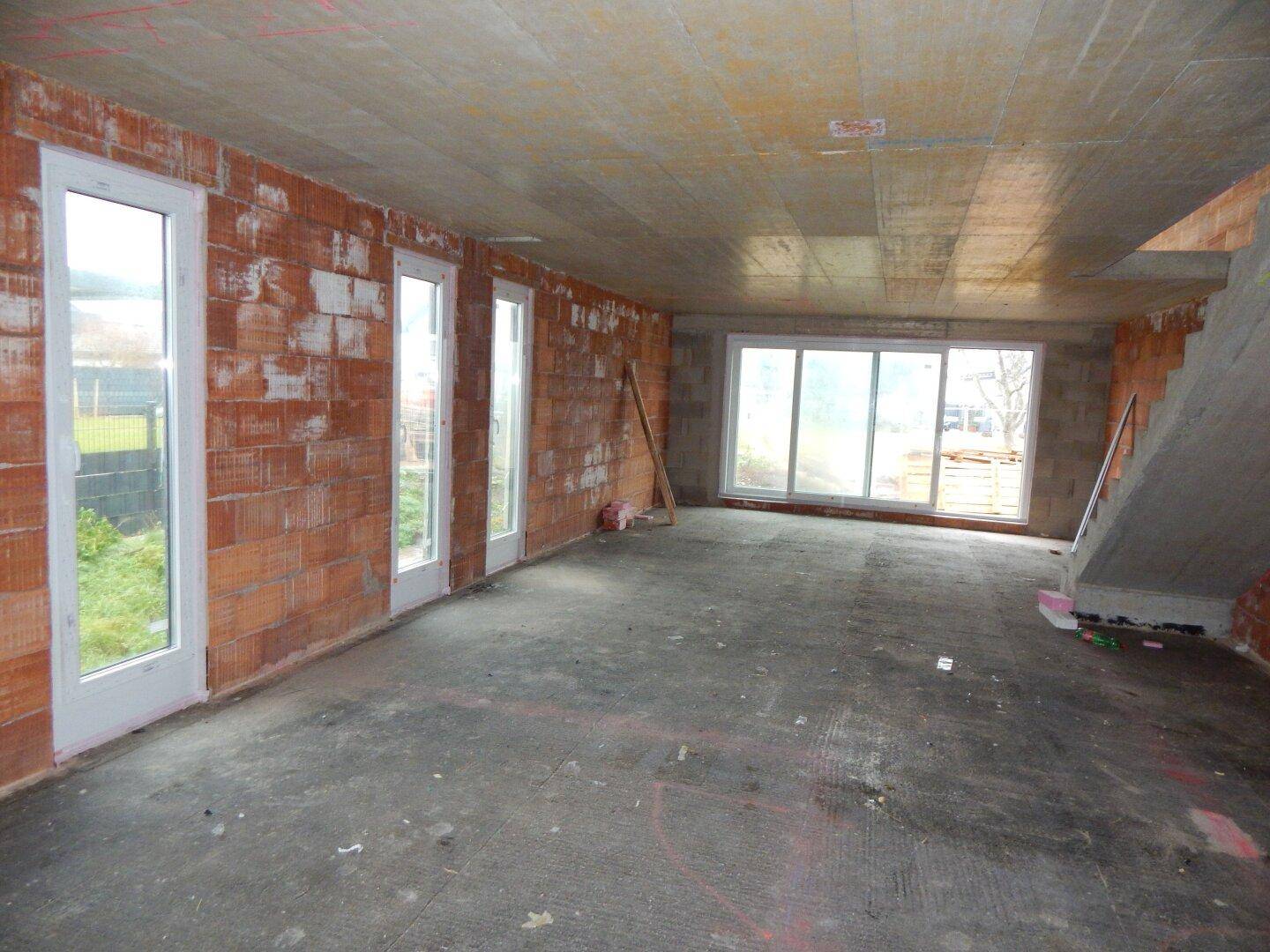 Rohbau eines hellen Wohnraums mit großen Fenstern und Zugang zum Garten, inklusive Treppenaufgang.