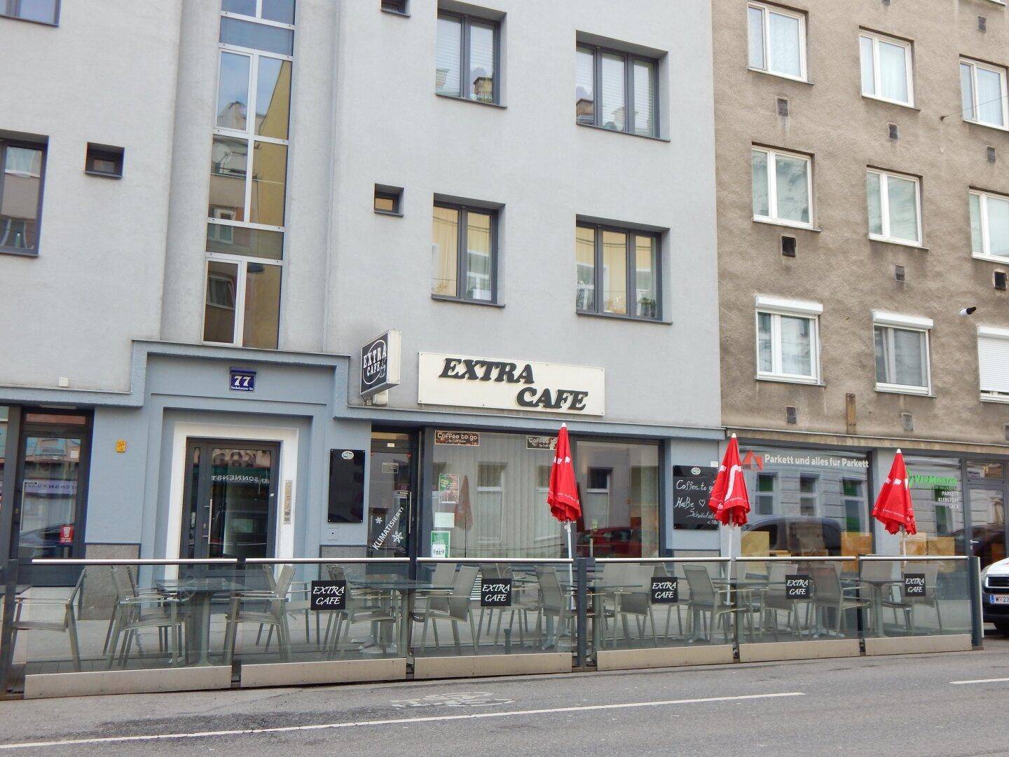 Gebäudeansicht mit Café und Bäckerei im Erdgeschoss, davor eine leere Terrasse.