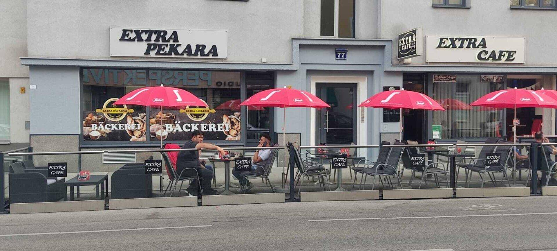 Straßencafé mit roten Sonnenschirmen und Glasabtrennung vor einer Bäckerei.