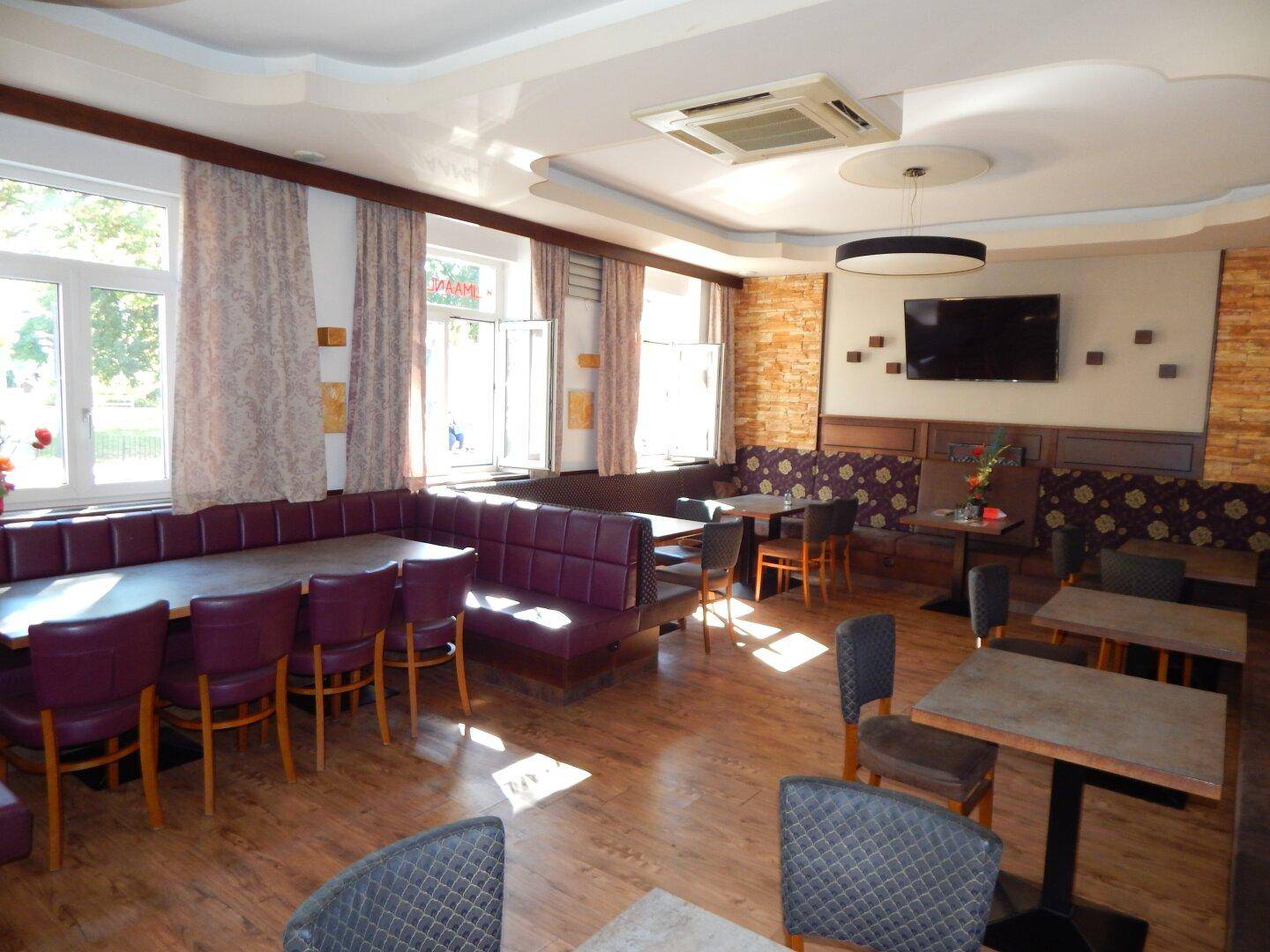 Heller Restaurantraum mit Holzboden, gepolsterten Sitzbänken und Tischen, beleuchtet durch große Fenster.